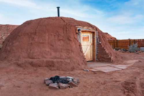 A traditional adobe structure with a wooden door and steps, set in a desert landscape with a fire pit nearby.